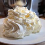 A generous swirl of light, fluffy vanilla buttercream frosting piped onto a white plate, with a mixer visible in the background.