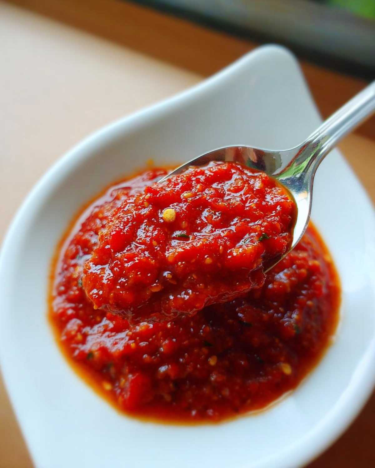 Close-up of thick, vibrant red pizza sauce being lifted from a white bowl with a metal spoon.