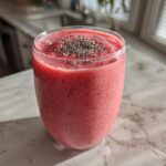 A vibrant pink watermelon smoothie in a glass, topped with chia seeds, sitting on a light countertop.