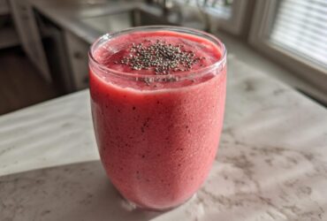 A vibrant pink watermelon smoothie in a glass, topped with chia seeds, sitting on a light countertop.