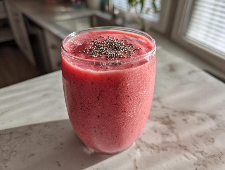 A vibrant pink watermelon smoothie in a glass, topped with chia seeds, sitting on a light countertop.