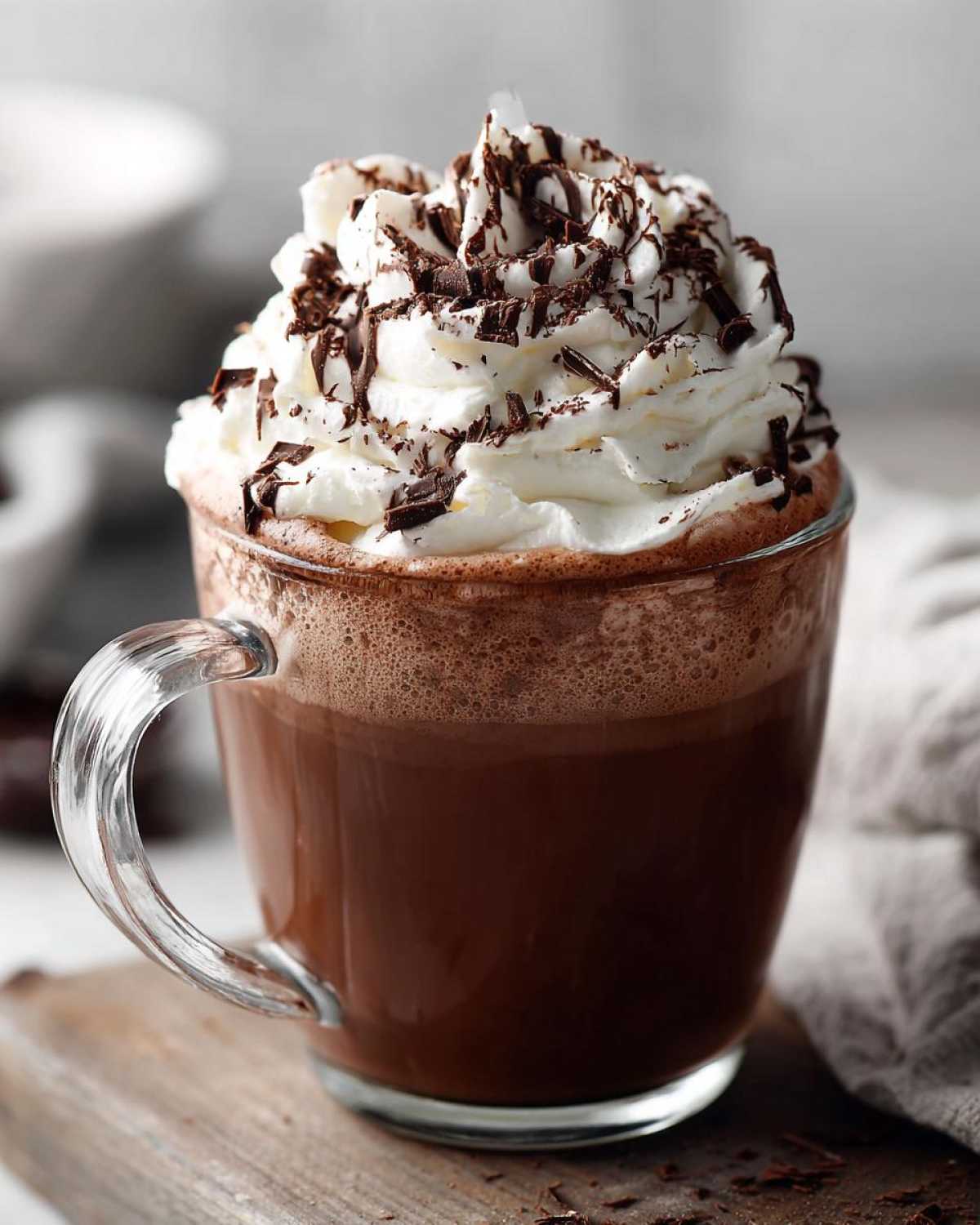 A clear glass mug filled with rich whipped hot chocolate topped high with whipped cream and dark chocolate shavings.
