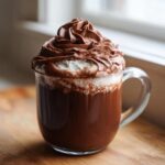 A clear glass mug filled with rich hot chocolate topped with white cream and decadent chocolate whipped hot chocolate.