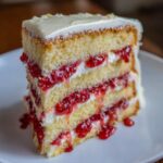 Close-up of a tall slice of white chocolate raspberry cake showing three layers of yellow cake, white frosting, and bright red raspberry filling.