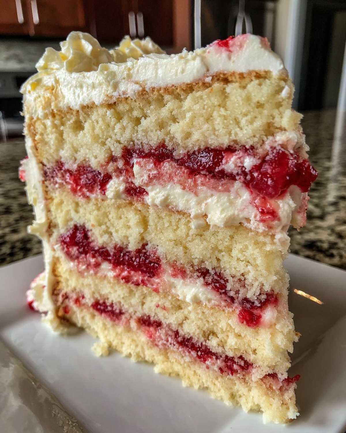 Close-up of a tall slice of white chocolate raspberry cake showing four layers of white cake, raspberry filling, and white frosting.