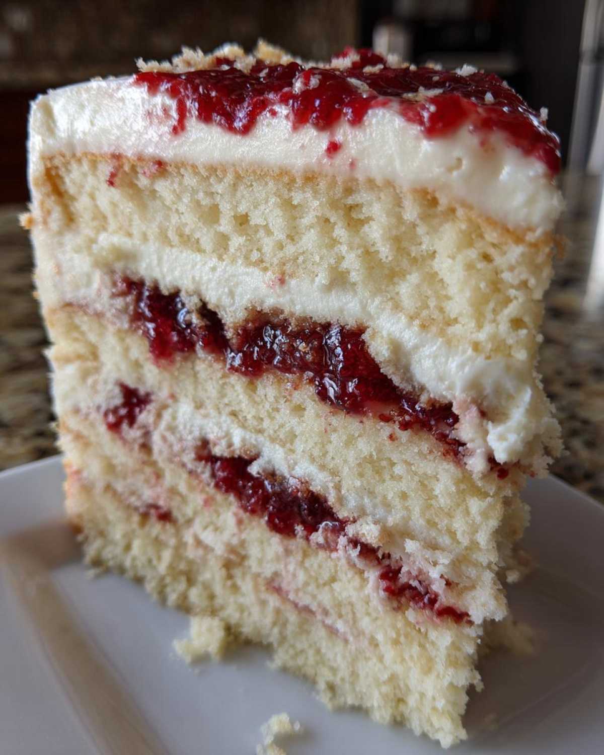 Close-up of a tall slice of white chocolate raspberry cake showing layers of white cake, cream, and bright red raspberry filling.