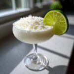 A creamy white Christmas margarita served in a coupe glass, topped with shredded coconut and garnished with a lime wheel.
