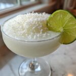 A frosty white Christmas margarita served in a coupe glass, topped with shredded coconut and garnished with a lime wheel.