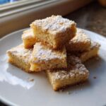 A stack of buttery, square shortbread cookies bars dusted heavily with powdered sugar on a white plate.