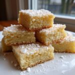 A stack of buttery, golden 3-ingredient shortbread cookies squares dusted generously with powdered sugar on a white plate.
