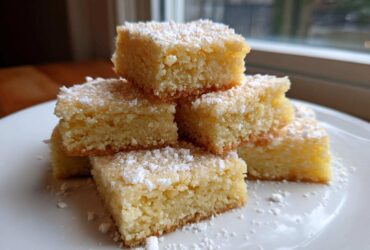 A stack of buttery, golden 3-ingredient shortbread cookies squares dusted generously with powdered sugar on a white plate.