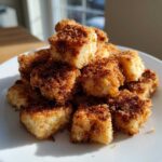A mound of golden Air Fryer Crispy Cinnamon Sugar Biscuit Bites coated heavily in cinnamon sugar on a white plate.