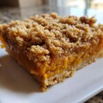 A close-up shot of a thick, square slice of pumpkin crisp bar with a gooey orange filling and crumbly topping.