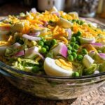 Close-up of a beautiful seven layer salad topped with shredded cheddar, hard-boiled eggs, peas, and red onion.