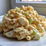 A generous scoop of creamy Amish macaroni salad featuring elbow pasta, celery, and hard-boiled egg chunks, served on a white plate.