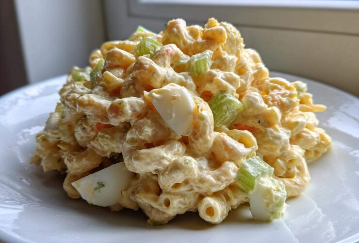 A generous scoop of creamy Amish macaroni salad featuring elbow pasta, celery, and hard-boiled egg chunks, served on a white plate.
