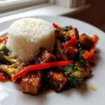 A plate featuring an Asian inspired Tofu Dish With Rice and Vegetables, glazed tofu cubes, broccoli, and red peppers.