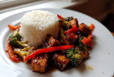 A plate featuring an Asian inspired Tofu Dish With Rice and Vegetables, glazed tofu cubes, broccoli, and red peppers.