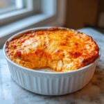 A baked cottage cheese buffalo chicken dip in a white round dish, showing a scoop removed and a melted, bubbly orange cheese topping.