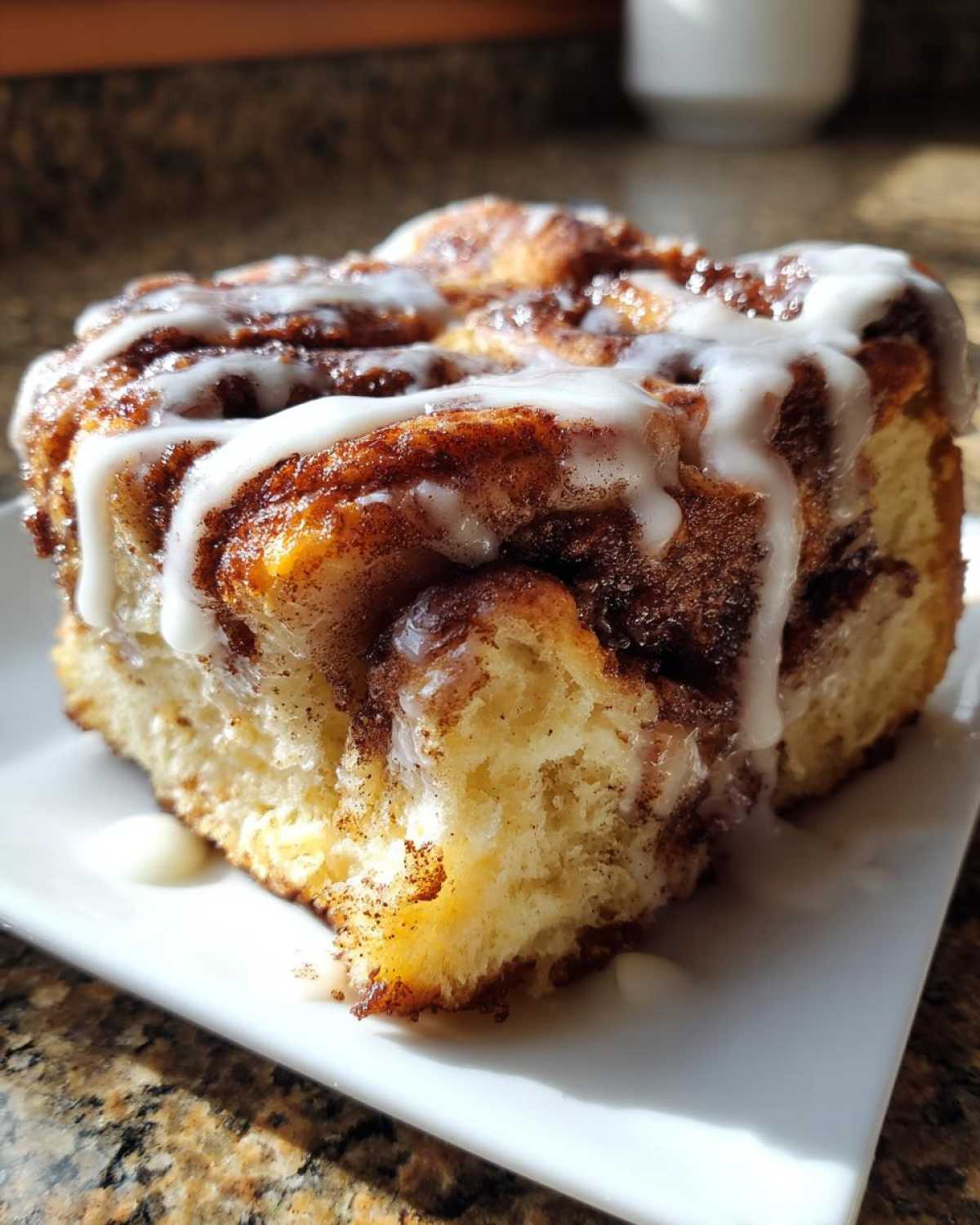 A single, fluffy serving of Banana Bread Batter Cinnamon Buns drizzled generously with white icing on a white plate.