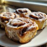 Four freshly baked Banana Bread Cinnamon Rolls with white icing drizzled on top, sitting on a white plate.
