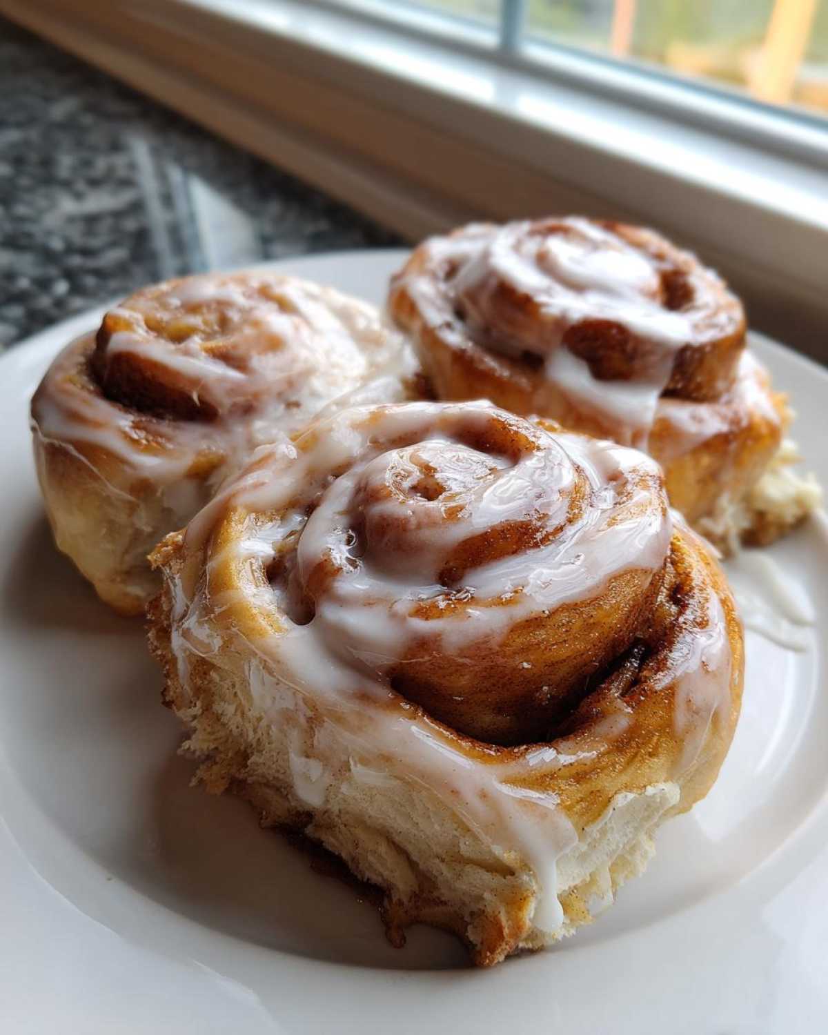 Three freshly baked Banana Bread Cinnamon Rolls Overnight Baking covered in thick white icing on a white plate.