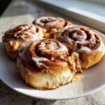 Four freshly baked Banana Bread Cinnamon Rolls drizzled with white icing on a white plate.