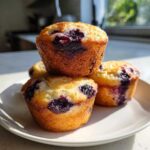 A stack of three golden-brown Blueberry Cottage Cheese Muffins bursting with dark purple berries, sitting on a white plate.