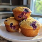 Three golden-brown Blueberry Cottage Cheese Muffins stacked on a white plate near a bright window.