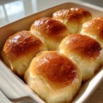 Close-up of six golden brown, glossy, buttery rolls baked closely together in a white dish.