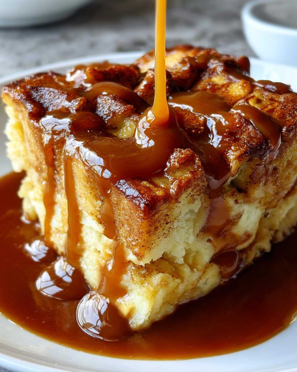 A close-up of a square serving of Cinnamon Bread Pudding being generously drizzled with rich caramel sauce.