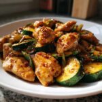 A close-up of a white plate filled with glistening chicken zucchini stir fry, topped with sesame seeds and chili flakes.