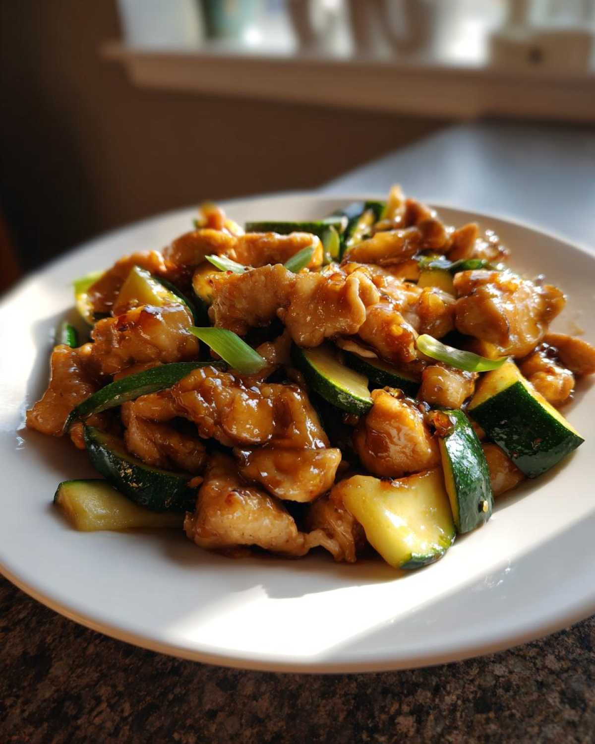 A plate featuring glistening pieces of chicken zucchini stir fry coated in a savory brown sauce, garnished with green onions.