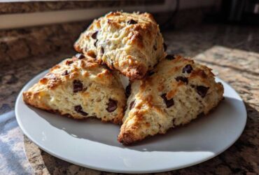 Three freshly baked chocolate chip scones, golden brown and studded with dark chocolate chunks, stacked on a white plate.