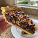 A close-up view of a rich slice of chocolate pecan pie being held up, showing the flaky crust and gooey filling.
