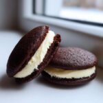 Two rich, dark chocolate whoopie pies filled with thick, creamy white frosting, sitting on a white surface.
