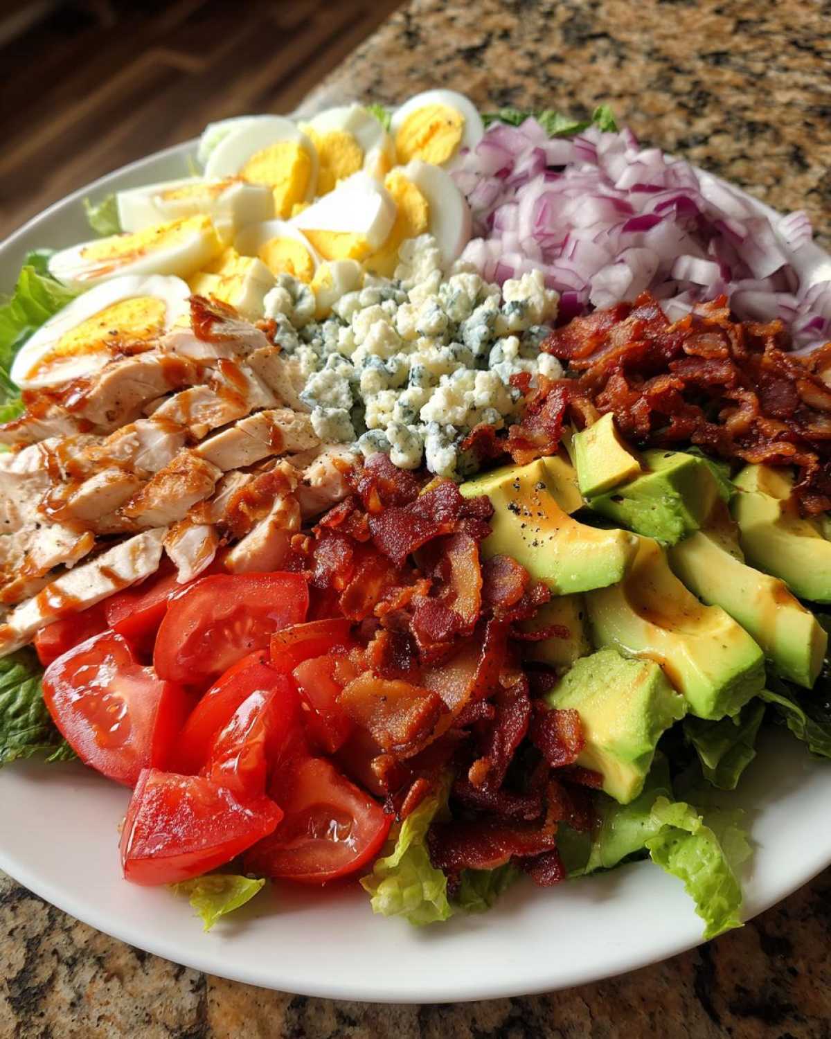 A beautifully arranged classic cobb salad featuring sliced chicken, hard-boiled eggs, blue cheese, bacon, avocado, tomatoes, and red onion.