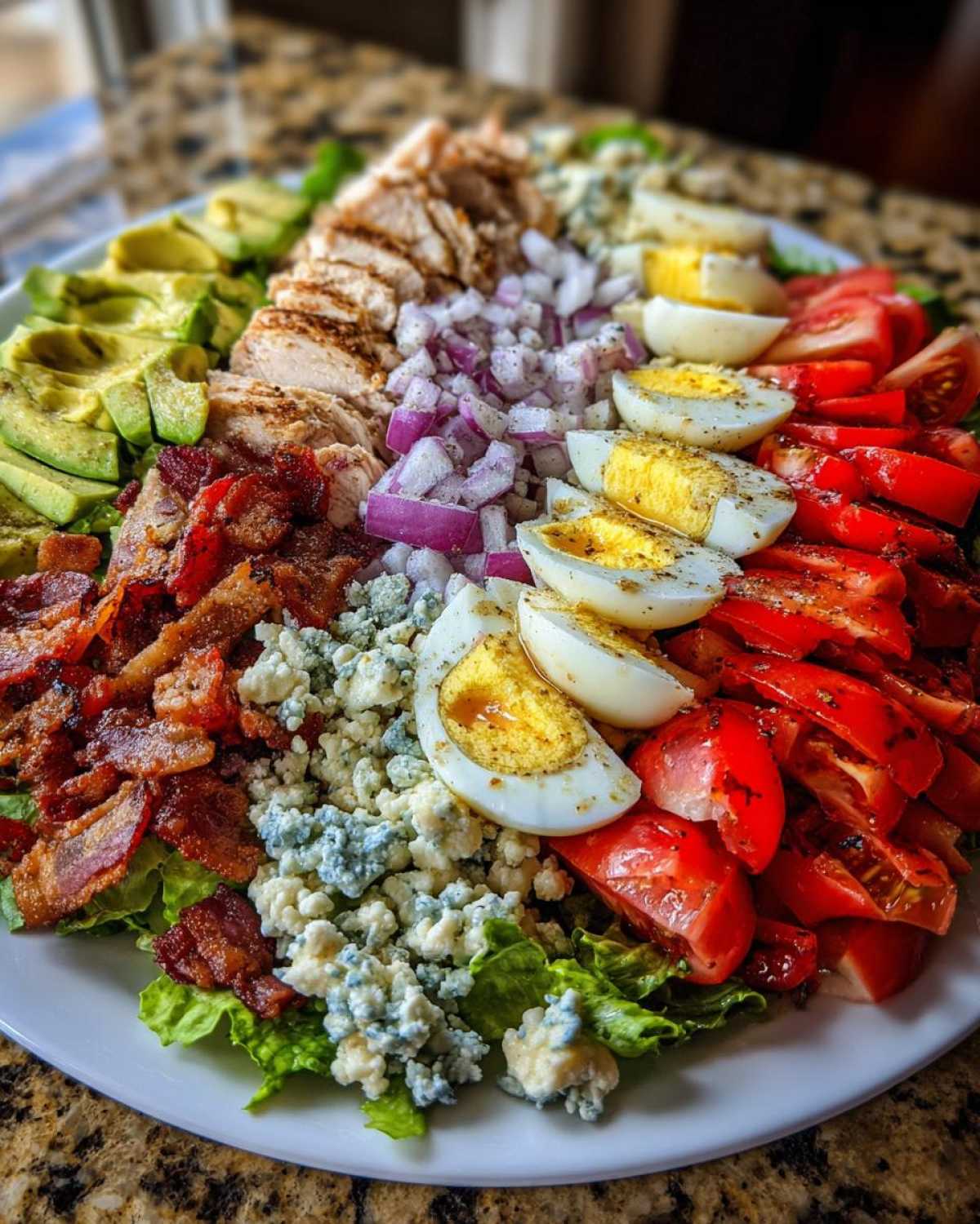 A vibrant, arranged cobb salad featuring rows of grilled chicken, avocado, bacon, blue cheese, eggs, and tomatoes.
