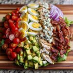 A vibrant, deconstructed cobb salad featuring rows of cherry tomatoes, hard-boiled eggs, avocado, chicken, blue cheese, bacon, and red onion.