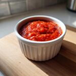 A small white ramekin filled with bright red, chunky cocktail sauce recipe, sitting on a wooden cutting board.