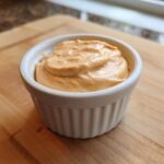 A small white ramekin filled with thick, orange-hued Copycat Classic Burger Spread Sauce sitting on a wooden cutting board.