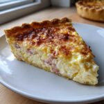 A close-up of a slice of You Should Really Try This Cottage Cheese Quiche on a white plate, showing a creamy filling with ham pieces.