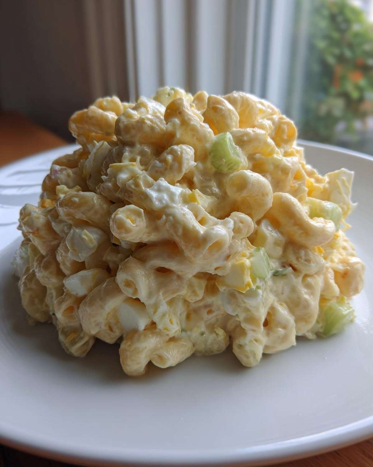 A generous scoop of creamy Amish macaroni salad featuring elbow macaroni, chopped egg, and celery on a white plate.