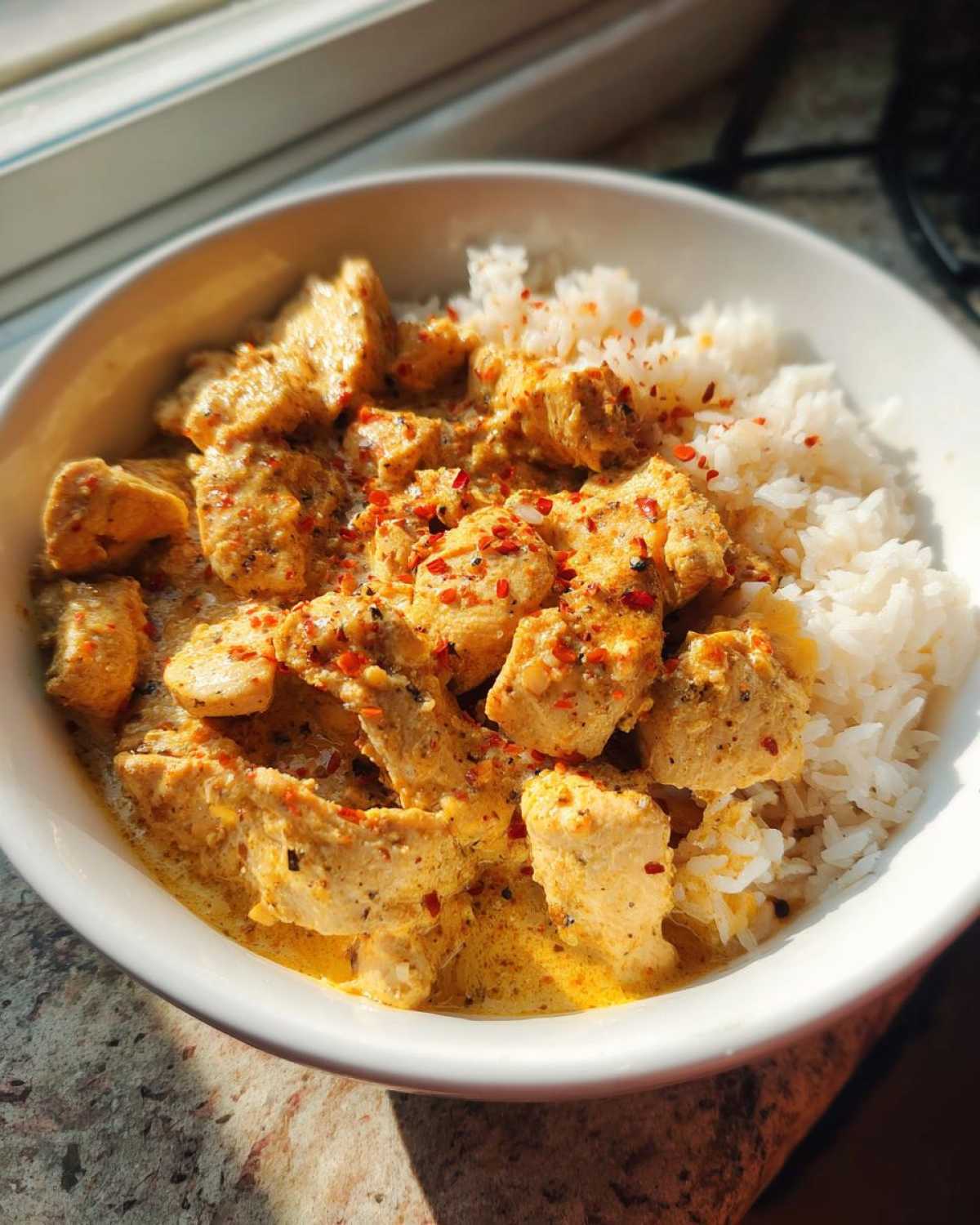 A white bowl filled with creamy cajun chicken pieces and fluffy white rice, topped with red pepper flakes.