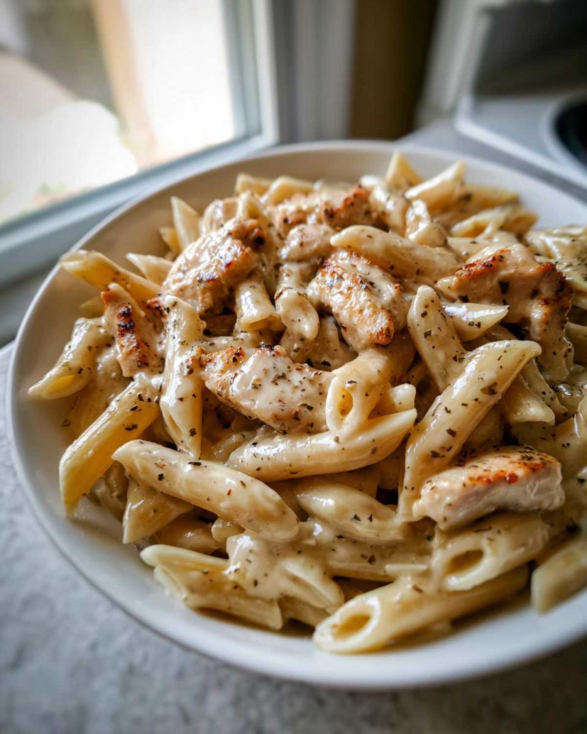 Close-up of creamy chicken pasta made with penne, topped with seasoned grilled chicken pieces in a white bowl.