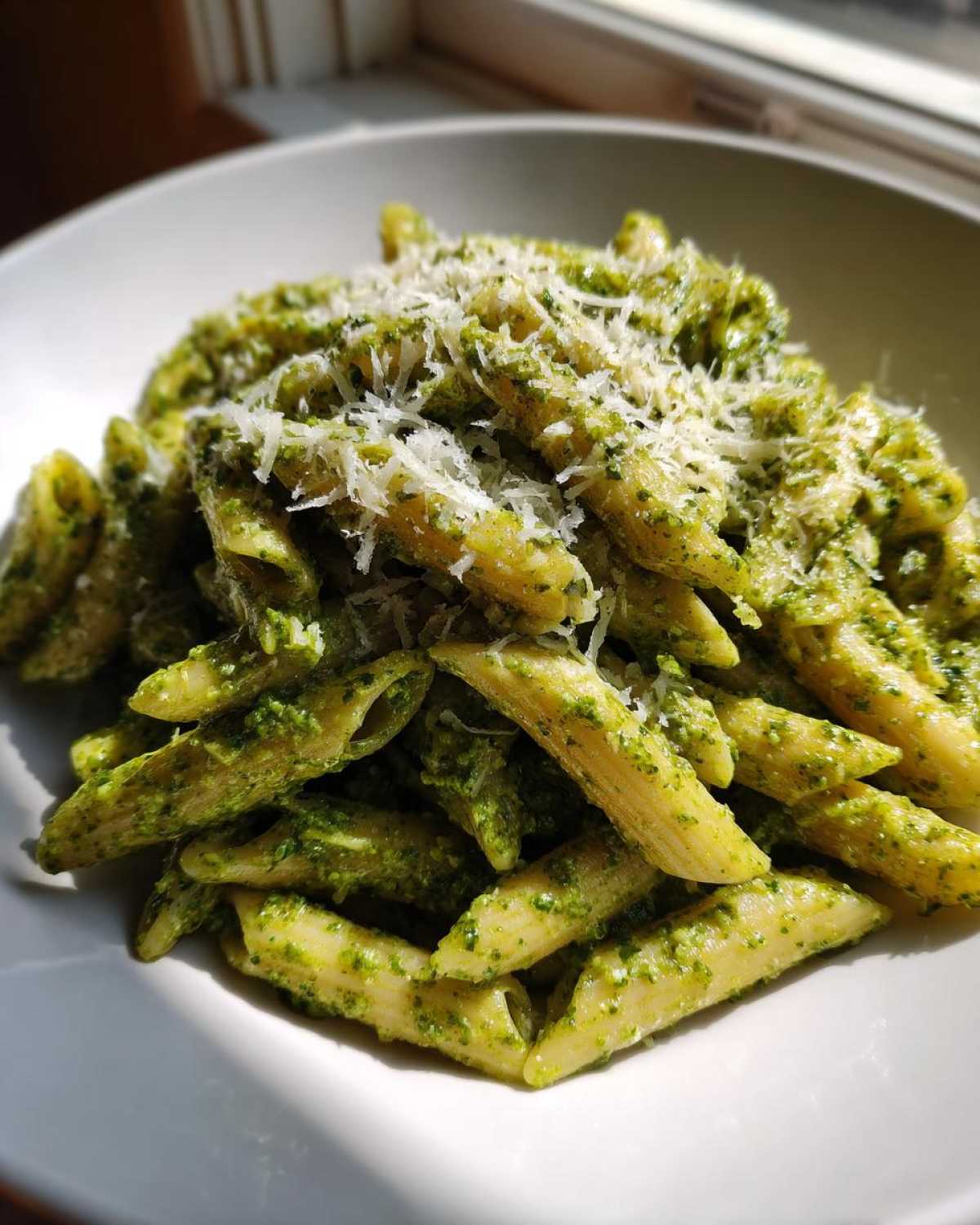 A close-up of penne pasta tossed in vibrant green sauce for creamy pesto pasta, topped with grated Parmesan cheese.