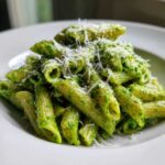 A close-up of penne pasta coated in vibrant green creamy pesto pasta, topped with freshly grated Parmesan cheese.