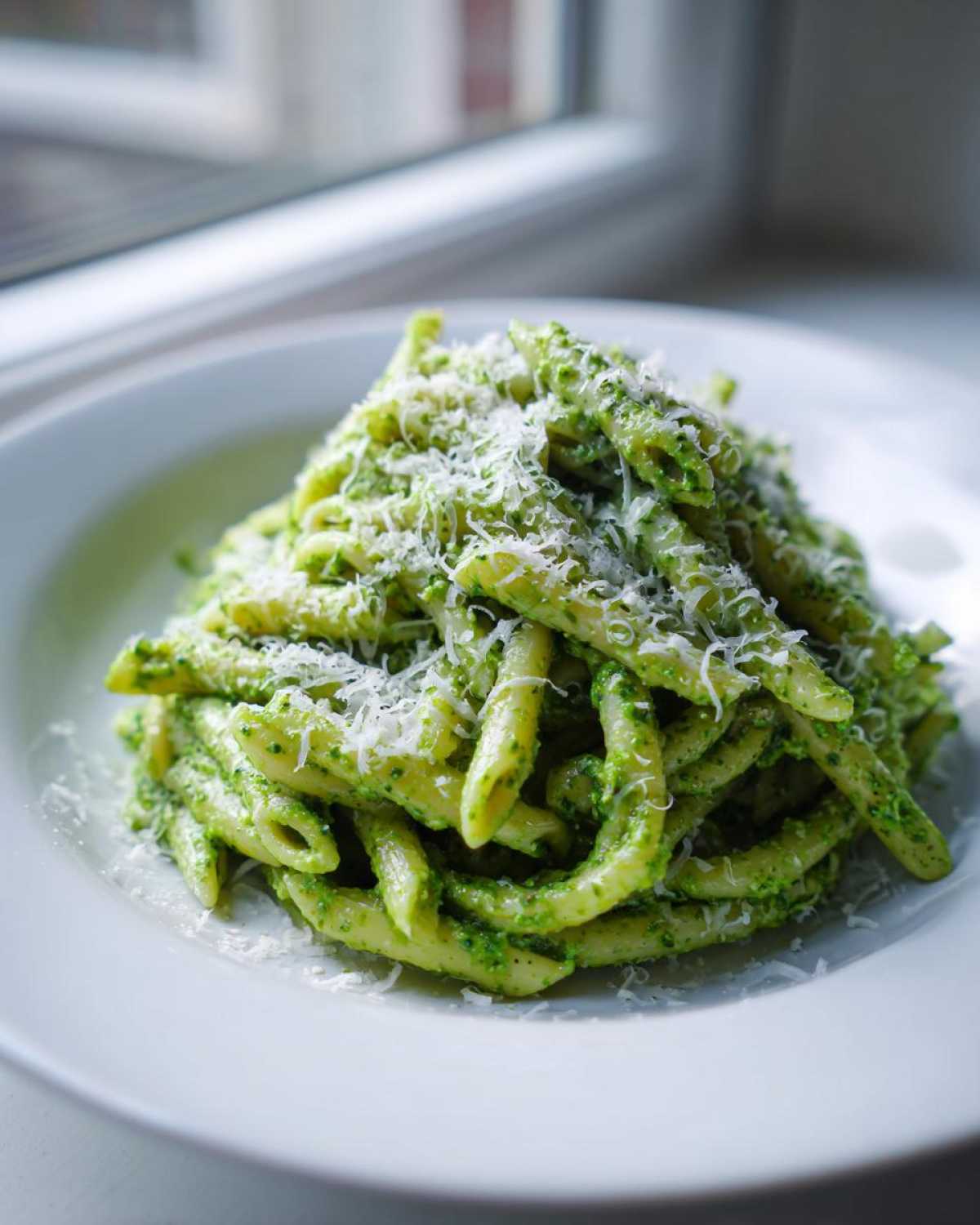 A close-up of a white plate piled high with creamy pesto pasta (penne) topped generously with grated Parmesan cheese.