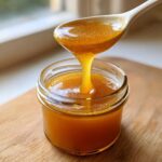 Close-up of thick, golden Dandelion Honey Syrup dripping from a white spoon back into a small glass jar.
