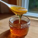 A white spoon drips thick, golden Dandelion Honey Syrup back into a small glass jar.